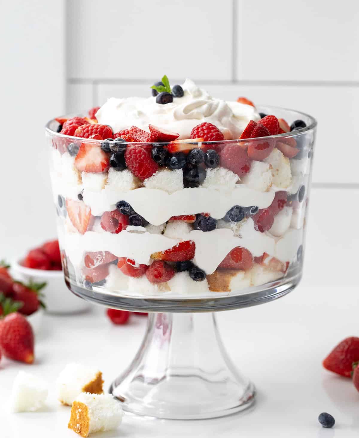 This Very Berry Trifle layers fluffy angel food cake, fresh strawberries and blueberries, and vanilla-orange whipped cream! Very Berry Trifle dish on a white counter with fresh fruit like strawberries and blueberries around the base.