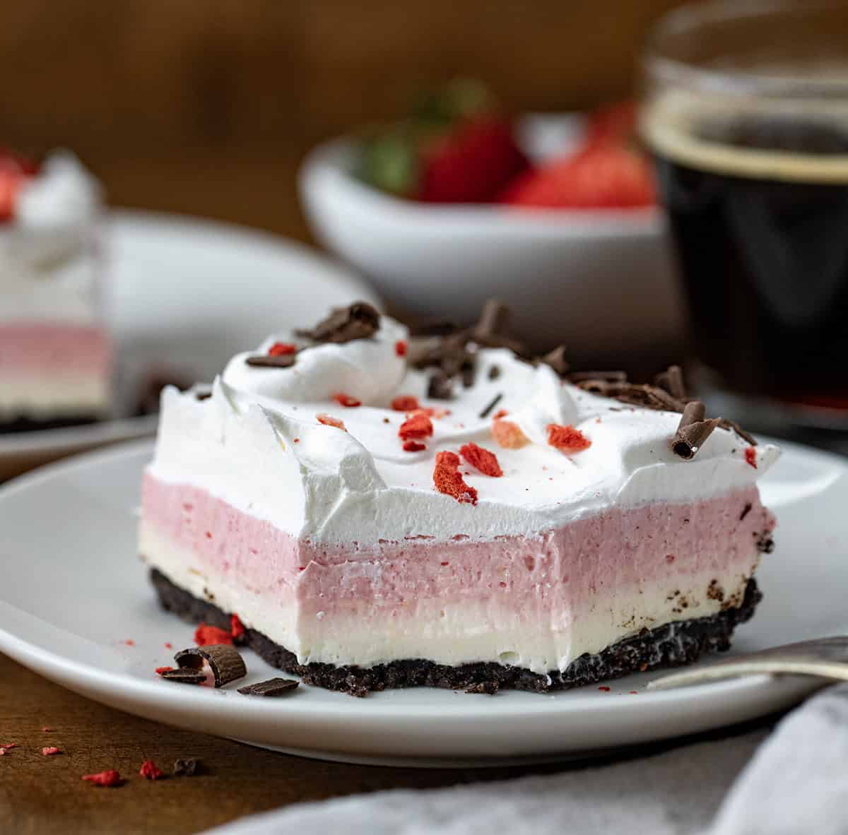 This Neapolitan dessert is a delicious layered no-bake treat that combines the classic flavors of chocolate, vanilla, and strawberry, like Neapolitan ice cream! It starts with a crunchy Oreo cookie crust, layered with creamy vanilla and strawberry fillings, and finished with a fluffy whipped topping. Neapolitan Dessert on a white plate with a bite removed and a fork resting on the plate.