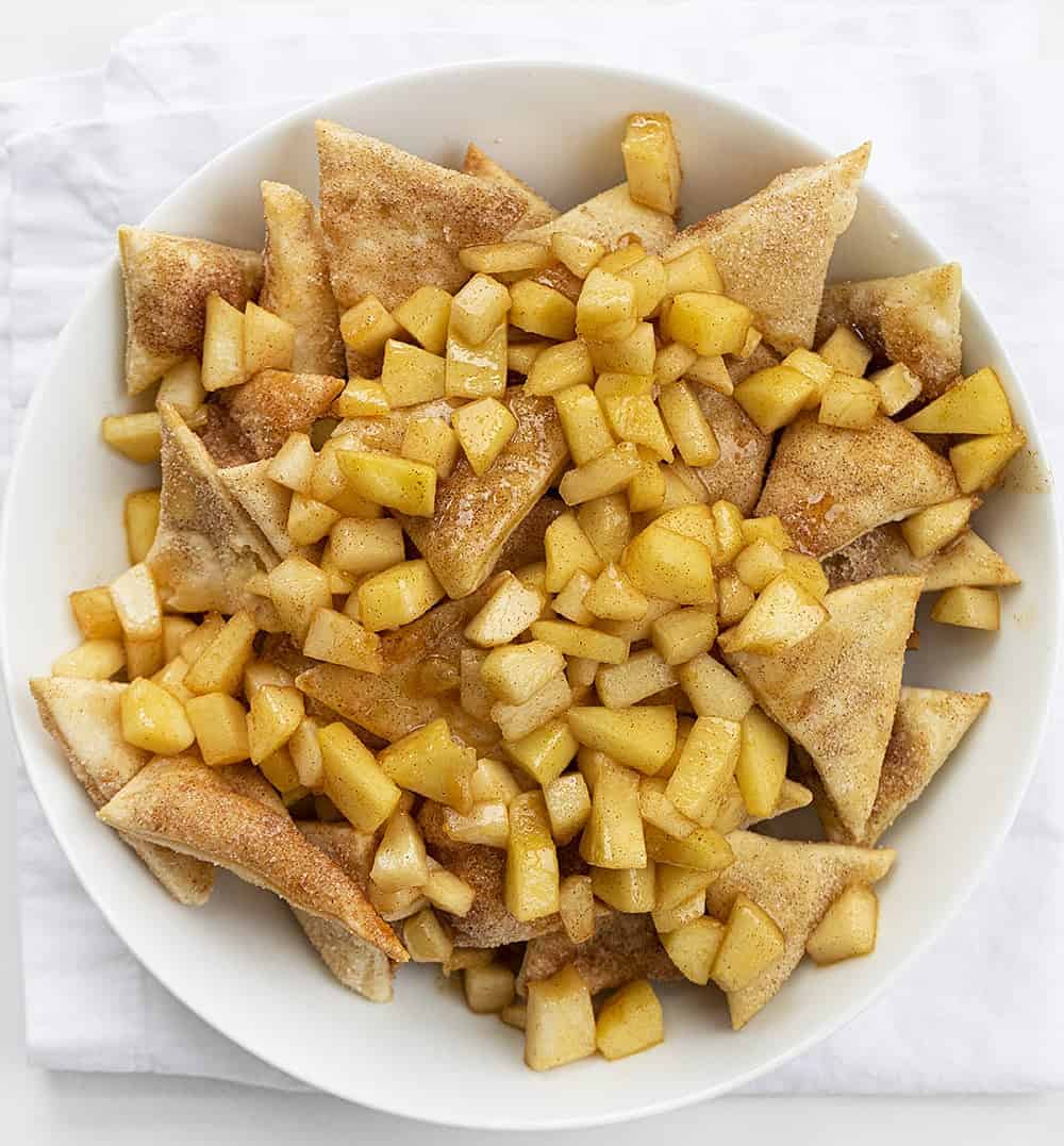 Apple Pie Dessert Nachos are homemade cinnamon-sugar chips, topped with sweetened apples, whipped cream, and drizzled with caramel. apple pie dessert nachos, dessert nachos, apple pie nachos, homemade whipped cream, baked cinnamon sugar chips, baking dessert recipes, apple pie, iambaker, i am baker Overhead Image of White Bowl with Homemade Apple Pie Nachos in it on White Napkin