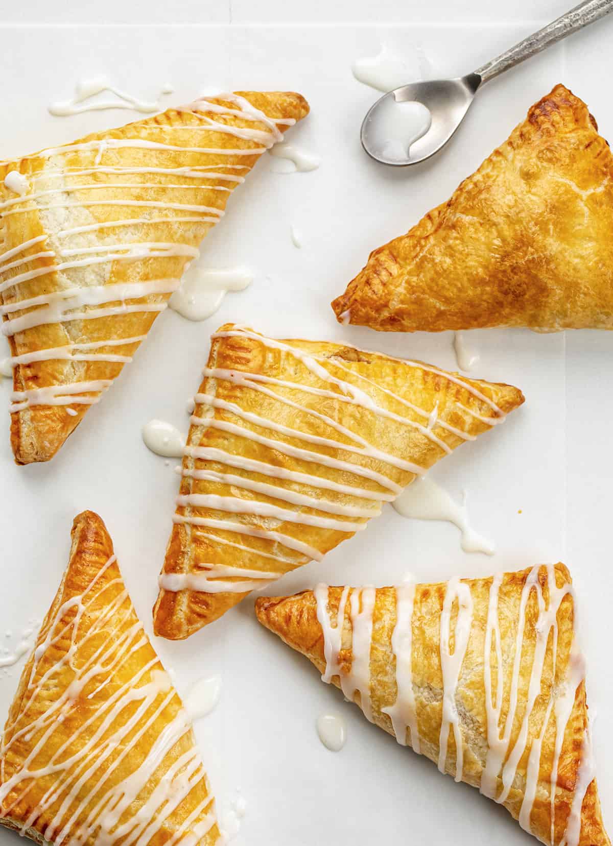 Easy & Amazing Apple Turnovers are filled with spiced apples and baked in flaky, buttery puff pastry dough! Apple Turnovers on a Counter with Glaze Drizzled Over Top and a Spoon.