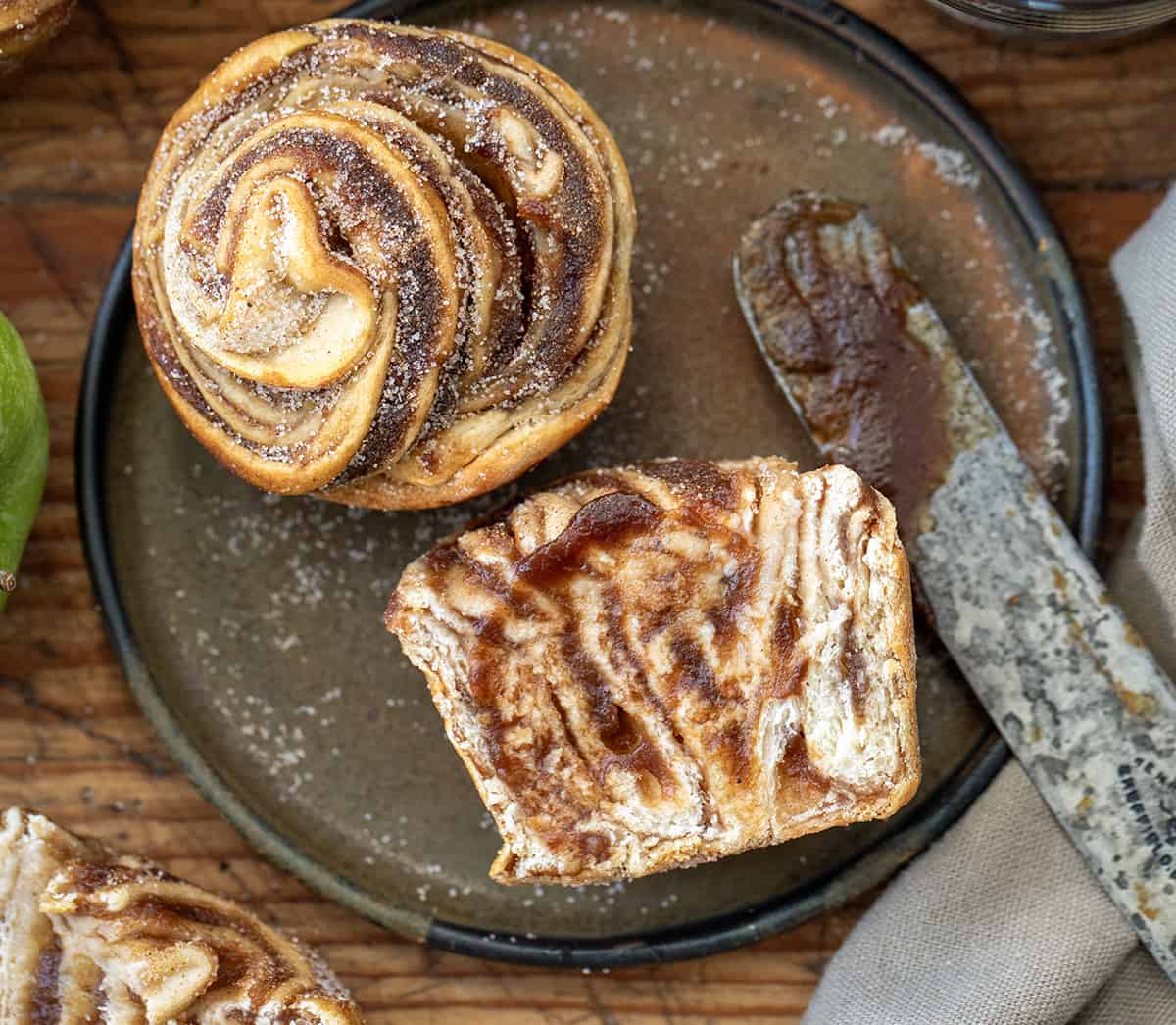 Apple Butter Cruffins are crescent roll dough smothered in homemade apple butter, rolled, cut, and baked in a muffin tin, topped with cinnamon and sugar. The entire recipe is made with just four ingredients! And, you get all the apple flavor without peeling or dicing any apples! Apple Butter Cruffins on a Plate with One Cut in Half and More Apple Butter.