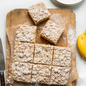 Banana Crumb Cake on a cutting board on a white table from overhead with some pieces cut and a banana on the side.