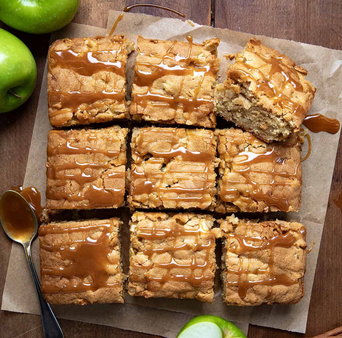This Caramel Apple Snack Cake is filled with fresh apples and flavored with apple pie spice, then baked and drizzled with caramel sauce. They are one of my favorite ways to use apples from the orchard in the Fall! Caramel Apple Snack Cake on a piece of parchment on a wooden table and cut into pieces.