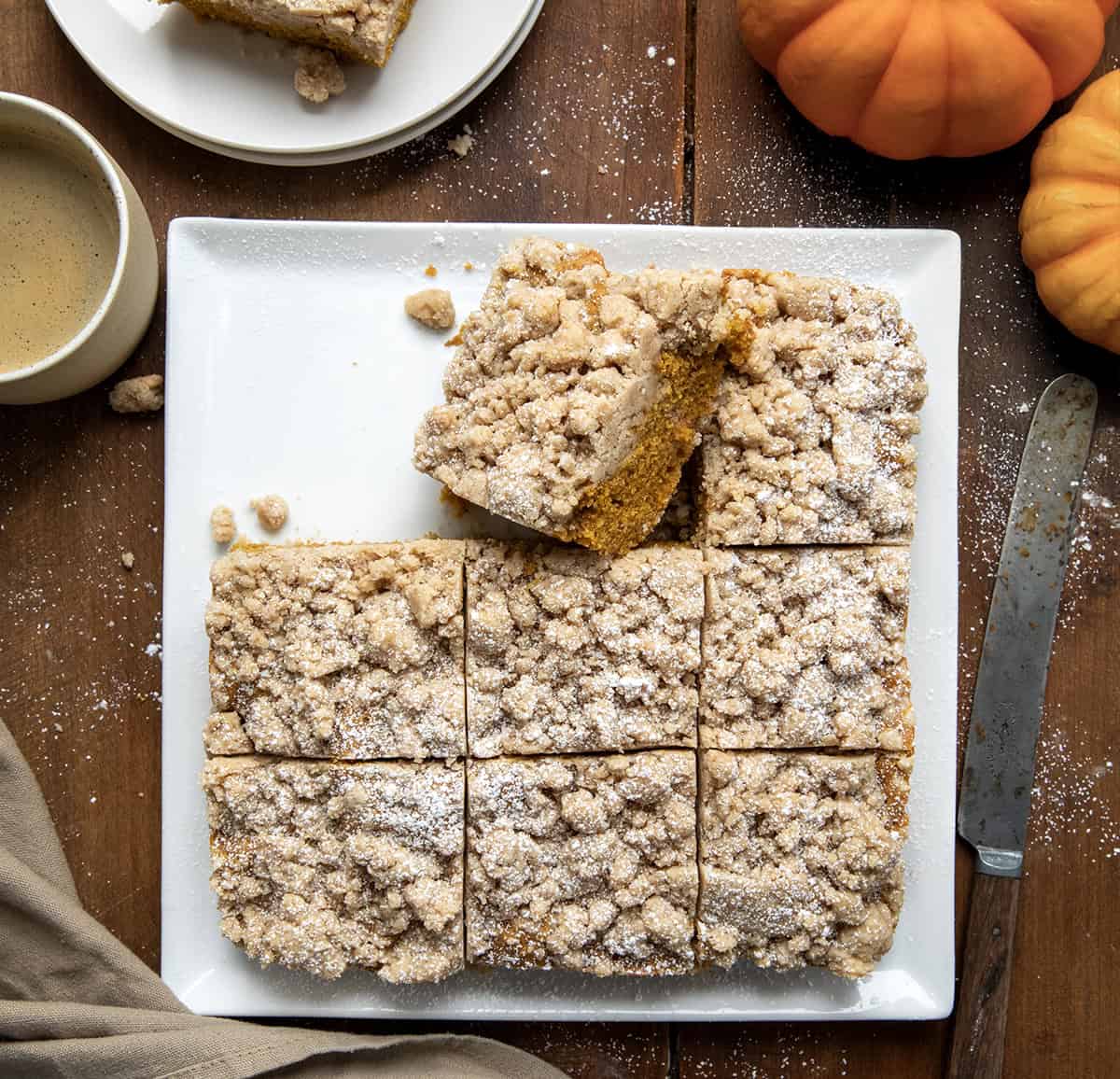 This Pumpkin Spice Coffee Cake is a moist, tender cake flavored with pure pumpkin and pumpkin pie spice, topped with a thick layer of crumbly streusel topping. These flavors are magical and will melt in your mouth! Pumpkin Spice Coffee Cake on a white platter cut into pieces with one piece on its side.
