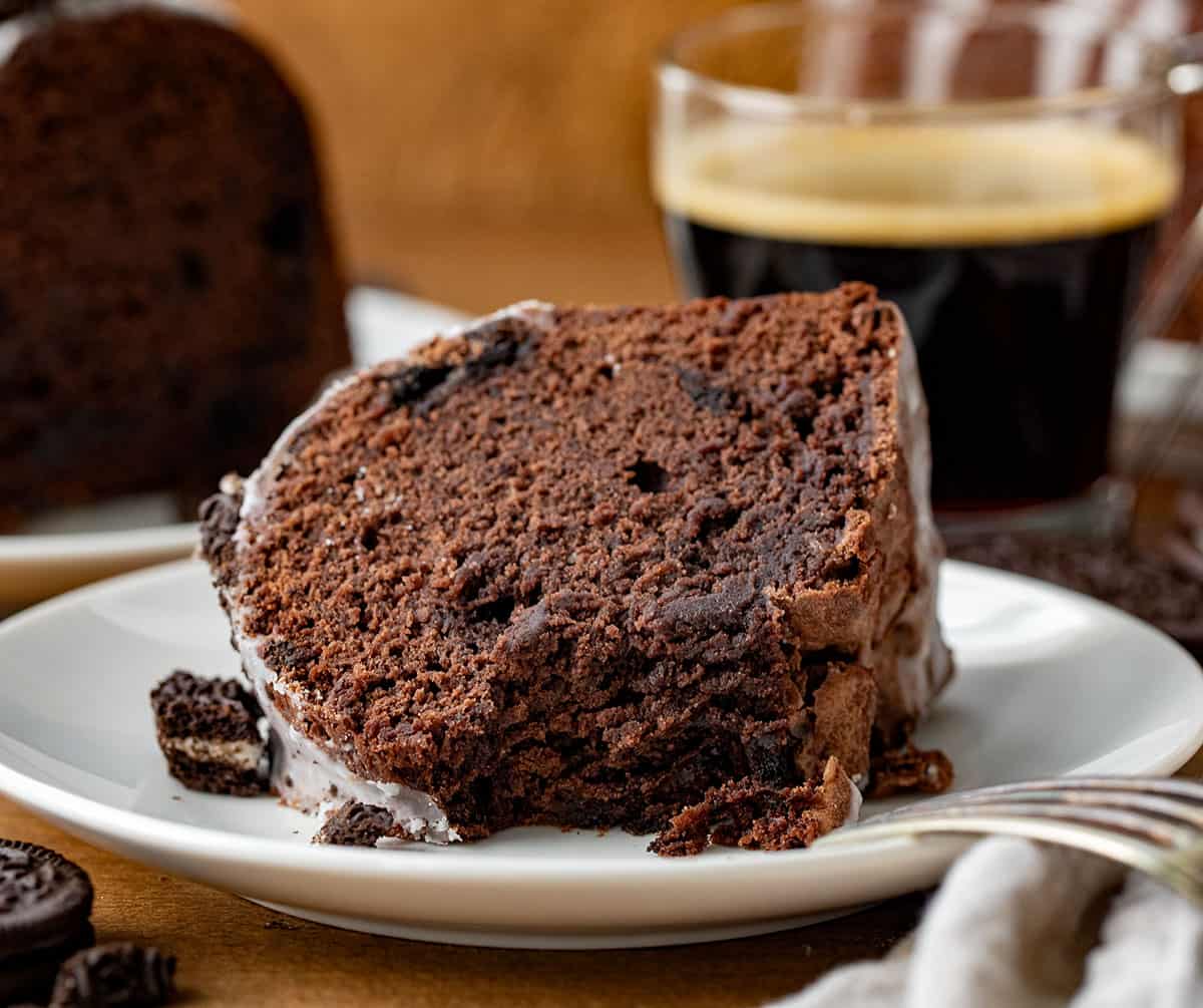 This Oreo Bundt Cake is a decadent take on my chocolate pound cake, packed with Oreo pieces, drizzled in a sweet glaze, and topped with more chopped Oreos. It's an Oreo lover's dream--perfectly blending chocolate, cream, and crunch in each slice! Piece of Oreo Bundt Cake on its side on a white plate with a bite taken out.